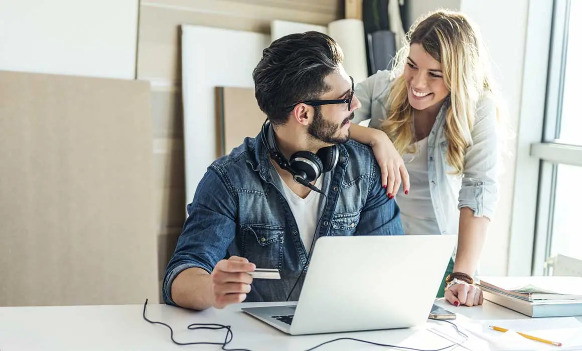 two people smiling at each other while one holds a credit card and sits in front of a computer